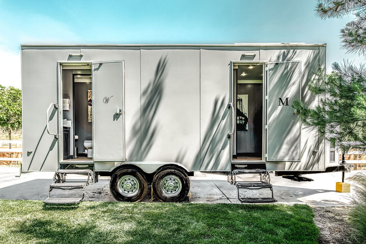 A luxury restroom trailer with both doors open, parked on a lawn in dappled afternoon light.